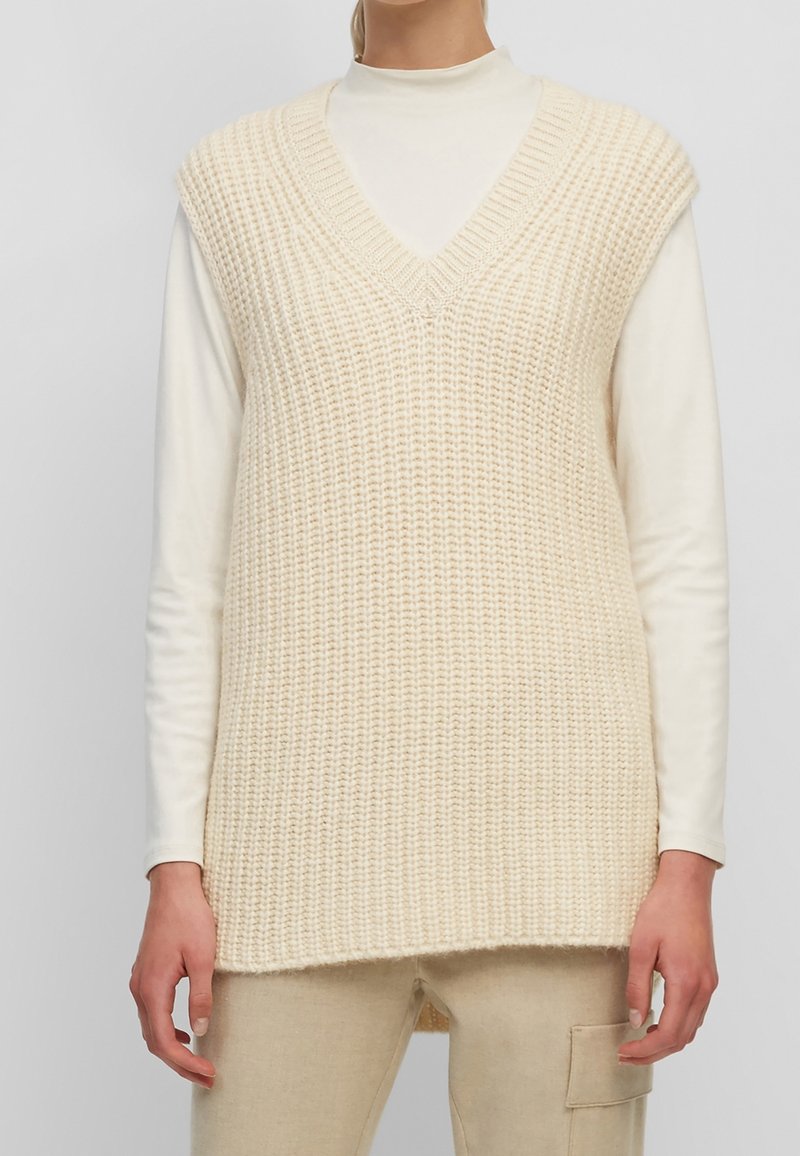 Gilet tricoté couleur crème avec col en V, motif texturé et design sans manches, superposé sur un haut blanc à manches longues.