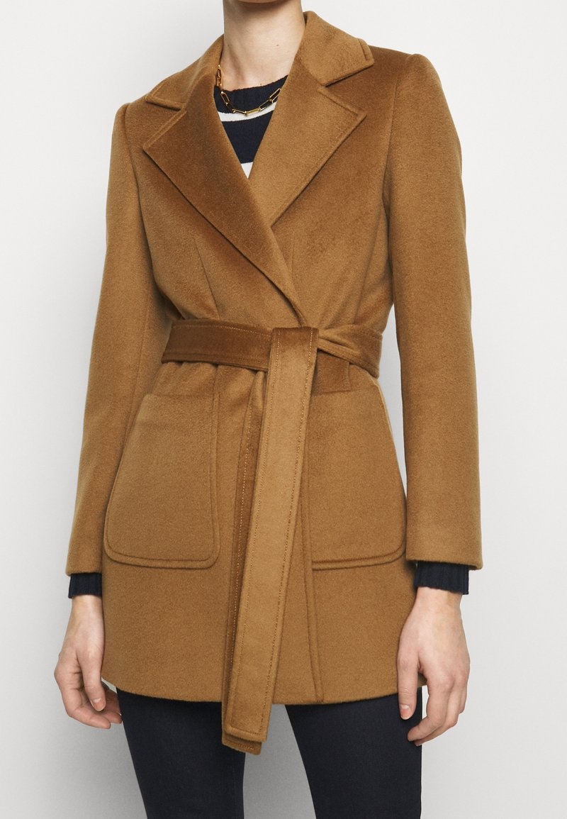 Mujer vistiendo un abrigo de lana color camel con cinturón, grandes solapas, bolsillos de parche y un suéter a rayas debajo.