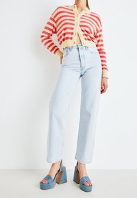 Proužkovaný cardigan v jemně žluté a růžové barvě, kombinovaný s světle modrými džíny s vysokým pasem a modrými platformovými šaty s dřevěnými podpatky a otevřenou špičkou.