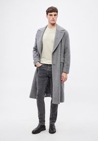 Γκρι παλτό με ψαροκόκαλο και φαρδύ γιακά, φορεμένο πάνω από ένα ανοιχτόχρωμο πουλόβερ, σε συνδυασμό με σκούρο τζιν και μαύρες μποτάκια μέχρι τον αστράγαλο.