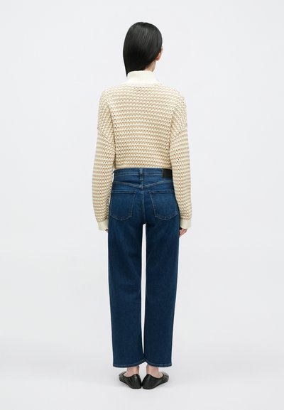 Camisola de malha creme com padrão texturizado, mangas largas e gola alta; combinada com jeans de ganga azul escura de cintura alta.