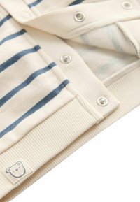 Krém színű babaruházat kék csíkokkal, patent gombokkal, bordázott mandzsettákkal és egy kis mackó logóval az alján. Puha anyag textúra.