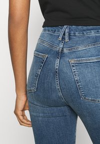 Högmidjade blå jeans i denim med en slät, texturerad yta, med två bakfickor och en dragsko i midjan.