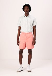 Lys grøn stribet kortærmet skjorte, ferskenfarvede shorts med elastisk talje, hvide sneakers og en sort bøttehat. Glat stof, afslappet pasform.
