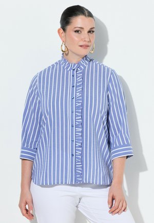 Kobieta z zaczesanymi do tyłu włosami, nosząca złote kolczyki-koła, niebiesko-białą koszulę w pionowe paski z falbankami oraz białe spodnie.