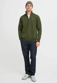 Olivengrønn hettegenser med halv glidelås og logo, kombinert med mørkeblå jeans og grå joggesko. Stoffet ser mykt og teksturert ut.