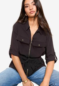 Melnā zipa krekls ar īsām ruļļveida piedurknēm, divām krūšu kabatām un saistītu jostas detaļu. Nēsāts ar ziliem džinsiem, akcentējot elegantu dizainu.