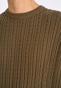 Brun stickad tröja med texturerade kabelmönster och rundad halsringning, tillverkad av ett mjukt tyg med vertikala ribbdetaljer.