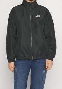 Svart vindjacka med hög krage, hel dragkedja fram, elastiska muddar och midjeband. Har en liten vit logotyp på vänster bröst.