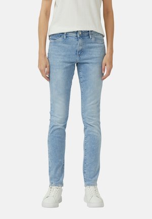 Personne portant un jean skinny bleu clair, des baskets blanches et un haut sans manches blanc, debout devant un fond uni.
