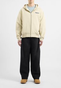 Felpa zip con cappuccio beige, con tasca e ricamo del logo, abbinata a pantaloni neri larghi e calzature beige. Tessuto liscio, vestibilità rilassata.