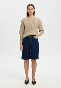 Suéter de punto beige, pantalones cortos de mezclilla azules, cinturón negro, zapatos marrones. El modelo está de pie contra un fondo neutro, con las manos en los bolsillos.