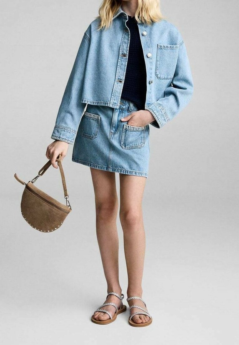 Femme portant une veste en jean bleu clair et une jupe assortie, un haut noir, tenant un sac à main marron en forme de demi-lune et portant des sandales argentées à brides.