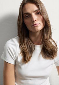 Top blanco de manga corta acanalado que lleva una persona con cabello largo y ondulado de color castaño. Textura suave y diseño minimalista, fondo neutro.