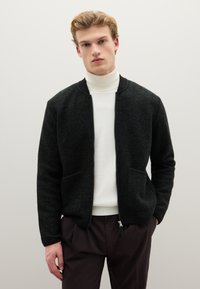 Bomber nero in pile con zip frontale, due tasche e polsini e collo a coste, indossato sopra un dolcevita bianco.