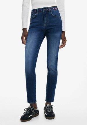 Jean taille haute bleu avec un léger délavage, accents floraux brodés à la taille, et coupe slim. Assorti à des baskets noires et grises.