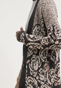 Czarny i beżowy kardigan z wzorem róż, wykonany z fakturowanej dzianiny, z zapięciem na guziki i kontrastującymi czarnymi rękawami.