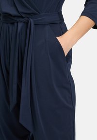 Tuta blu navy con design avvolgente, cintura in vita, maniche lunghe e tasche laterali, realizzata in tessuto morbido e elastico.