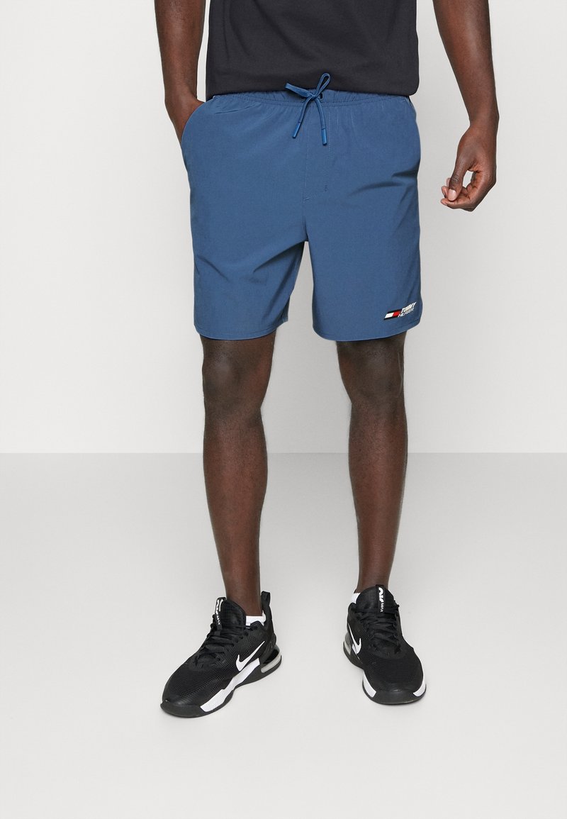 Pantalones cortos de natación elásticos azules con cintura ajustable con cordón, bolsillos laterales y un pequeño logotipo, combinados con zapatillas deportivas negras.