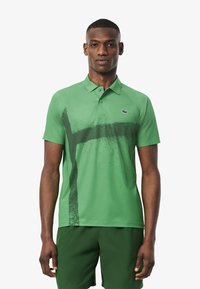 Grön polo-shirt med ett stänkmönster i mörkare grönt, korta ärmar och krage. Lacoste-logotyp på vänster bröst.