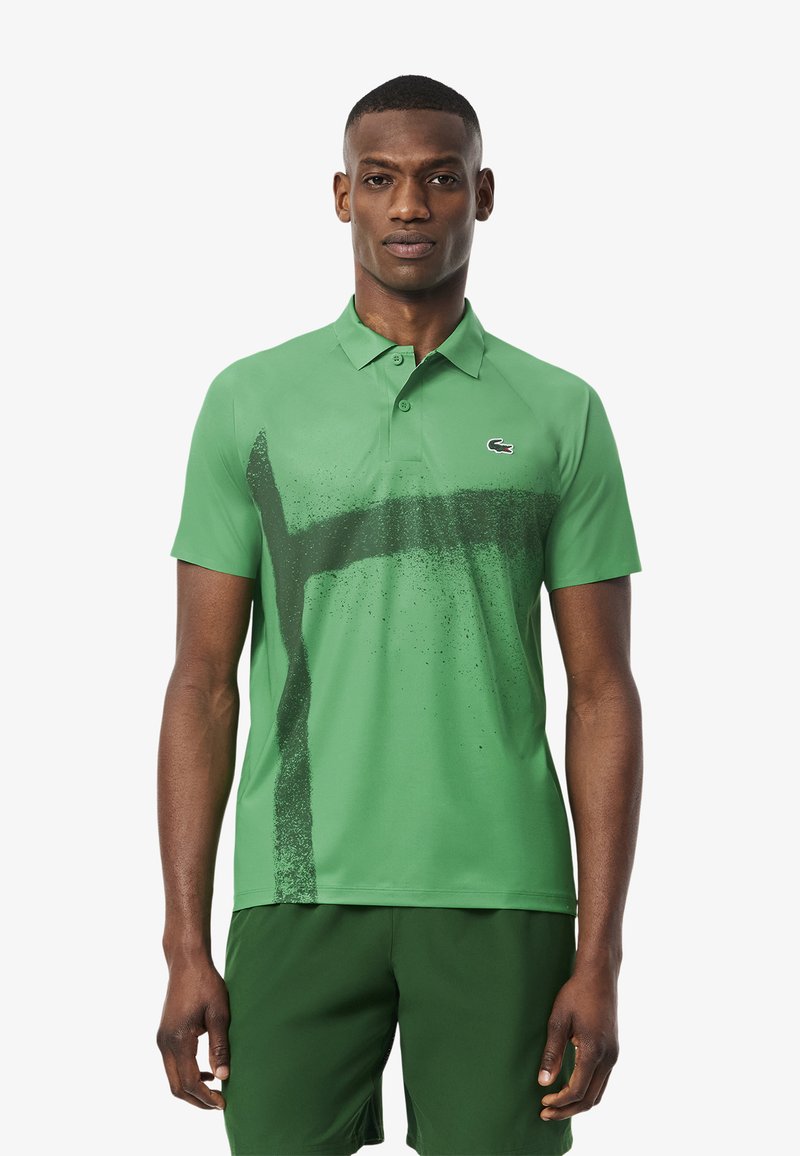 Grön polo-shirt med ett stänkmönster i mörkare grönt, korta ärmar och krage. Lacoste-logotyp på vänster bröst.