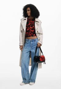 Femme aux cheveux bouclés porte un trench-coat beige, un haut floral rouge, un jean évasé, des chaussures blanches et tient un sac à main noir avec une grande fleur rouge.