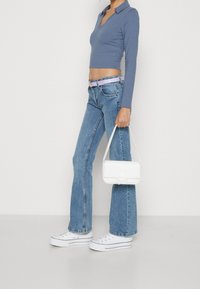 Kvinna i blå croppad långärmad tröja, högmidjade jeans med lavendelbälte, vita sneakers och bär en liten vit handväska.