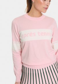 Vaaleanpunainen neule, jossa on valkoinen nauha, jossa lukee "après tennis." Pitkät hihat ribbaatuilla hihoilla. Sileä neulepinta.