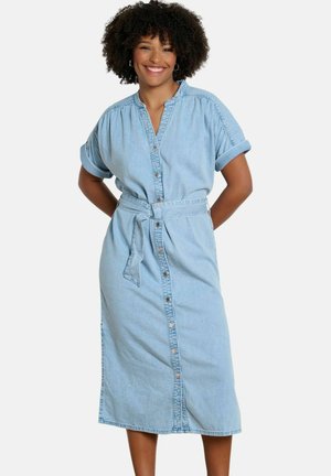 Šypsanti moteris su garbanotais plaukais, dėvinti šviesiai mėlyną džinsinę suknelę su sagutėmis priekyje, užriestomis trumpomis rankovėmis ir surišta juosmens diržu.