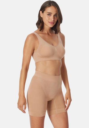 Vrouw die een naadloze sportbeha in naaktkleur en hooggetailleerde biker shorts modelleert, staand tegen een effen lichtgrijze achtergrond.