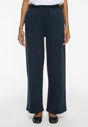 Personne portant un pantalon ample bleu marine foncé avec une ceinture élastique et des chaussures plates noires, debout devant un fond uni.