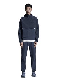 Veste de sport bleu marine avec fermeture éclair, capuche et bande réfléchissante, associée à un pantalon fuselé assorti. Le mannequin porte des baskets grises.