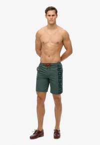 Grüne Badeshorts mit elastischem Bund, orangefarbenem Kordelzug und großem "SUPERDRY"-Druck an der Seite. Getragen mit braunen Schuhen.