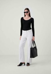 Donna che indossa una maglietta nera a maniche lunghe, pantaloni bianchi a gamba larga, tacchi aperti neri, occhiali da sole rettangolari di grandi dimensioni, mentre tiene una borsa a tracolla nera.