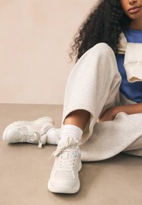 Gaiši pelēki sporta apavi ar tīkla un teksturētām panelēm, ar gaiši zilu akcentu un plakanām šņorēm. Kopā ar pelēkām sporta biksēm un baltām zeķēm.