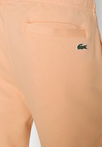 Persikofärgade mjukisbyxor i mjukt tyg, med en bakficka och en liten grön krokodillogo. Slät textur.