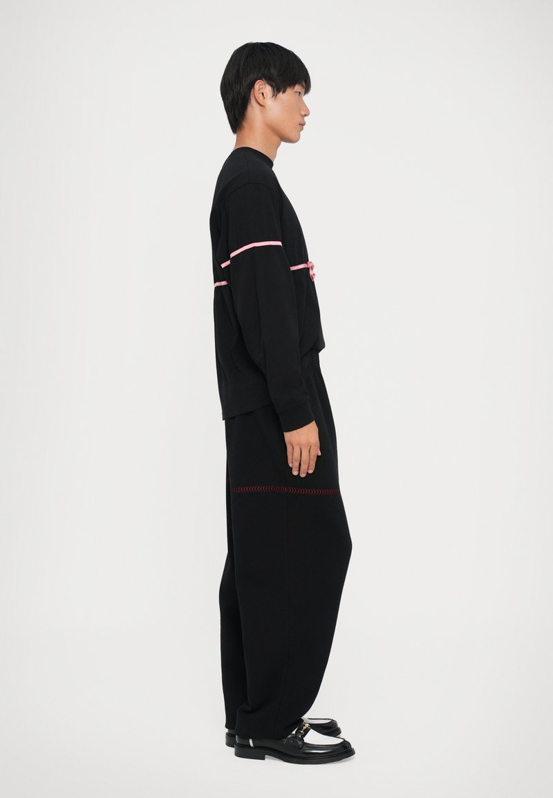 Camisa negra de manga larga con acentos de cinta rosa, combinada con pantalones negros holgados que tienen costuras rojas. El modelo lleva mocasines negros.