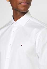 Camicia bianca con bottoni realizzata in cotone, con colletto classico e un piccolo logo ricamato in rosso, bianco e blu sulla parte sinistra del petto.