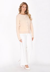 Cardigan beige chiaro con rifiniture bianche e chiusura con bottoni, indossato sopra pantaloni bianchi a gamba larga. Presenta maniche lunghe e orlo a coste.