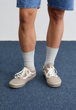 Persoon draagt beige sneakers met witte veters, witte enkelsokken en blauwe spijkershorts, staand op een blauw tapijt.