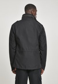 Black jacket with a high collar, made of smooth fabric. Features shoulder epaulets and a straight cut with a slightly longer back hem.