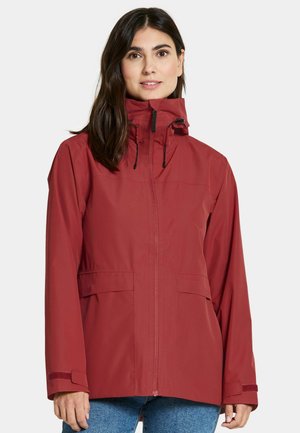 Frau trägt eine rote wasserdichte Jacke mit hohem Kragen, Frontreißverschluss und verstellbaren Manschetten, kombiniert mit blauen Jeans.