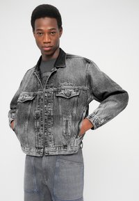 Džinsinė striukė išblukusiu juodu atspalviu su kontrastingu juodu apykakle, sagų užsegimu, dviem priekinėmis kišenėmis ir ant krūtinės siuvinėtu „Diesel Industry“ logotipu.