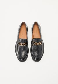 Sorte lær loafers med preget logo mønster, en glatt tekstur, og en gullkjede aksent over fronten.