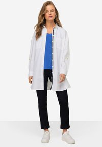 Camicia bianca a maniche lunghe con dettagli gialli sopra un top blu, indossata con jeans scuri e sneakers bianche. Vestibilità comoda.