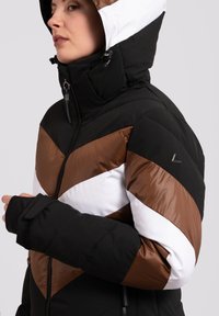 Puffermantel mit Chevron-Muster in Schwarz, Braun und Weiß. Verfügt über einen hohen Kragen, Reißverschlusstaschen und eine verstellbare Kapuze mit Befestigungen.