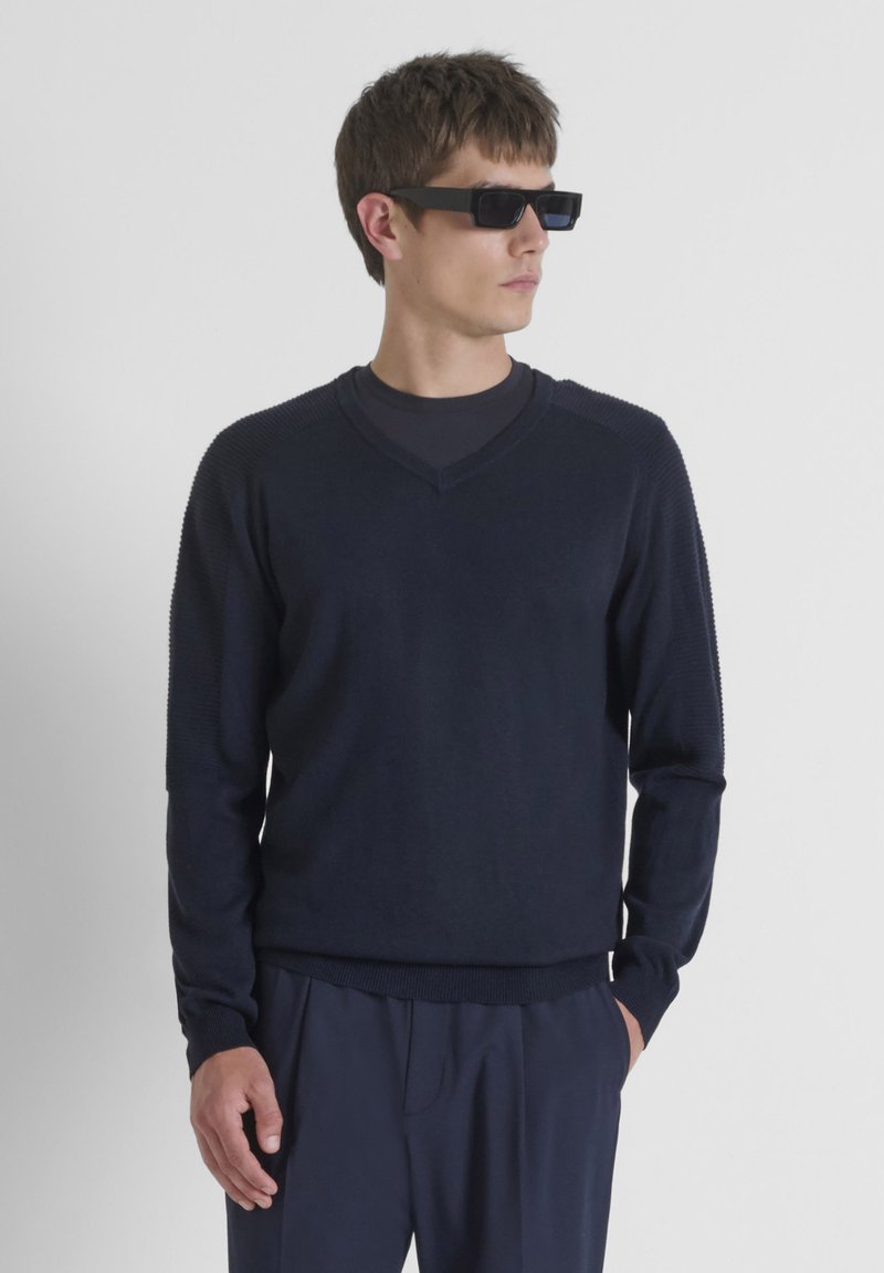 Navyblauwe gebreide trui met V-hals, geribbelde textuur op de schouders en aansluitende mouwen, gecombineerd met donkere broeken.