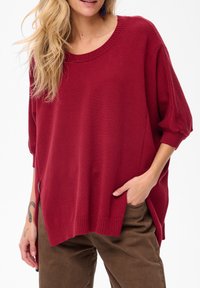 Kobieta ubrana w oversize'owy, czerwony sweter z włóczki, z rękawami do łokcia i brązowymi spodniami, z ręką w kieszeni, z tatuażem widocznym na lewym przedramieniu.