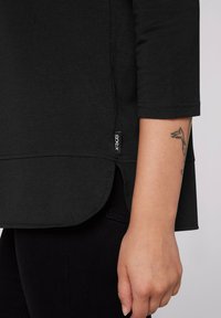 Camisa negra de manga larga con dobladillo redondeado y aberturas laterales, y una pequeña etiqueta con el logo en la parte inferior izquierda. La tela parece suave y lisa.