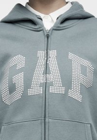Gros plan sur une personne portant un sweat à capuche gris zippé avec des lettres en strass formant le mot "GAP" sur une chemise blanche à col.